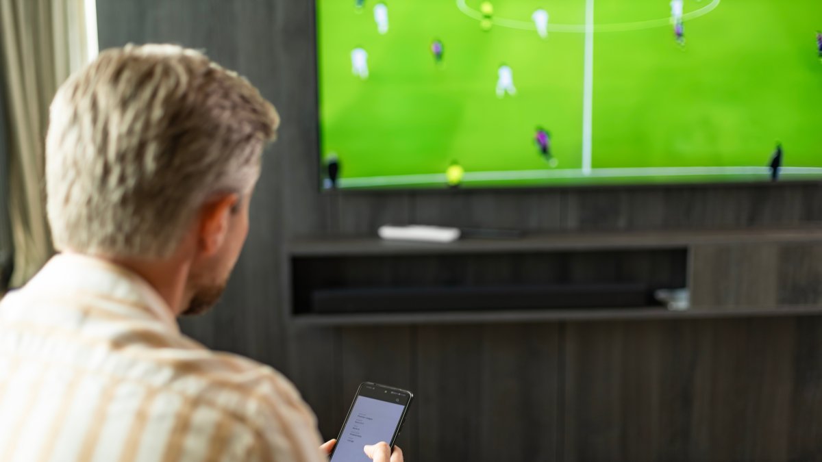 A man uses a smartphone betting app while watching a football match. (Shutterstock Photo)