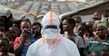 A health worker in personal protective equipment (PPE) stands near displaced people waiting for the burial of suspected Ebola victims at the Kigonze displaced persons camp, one month after an outbreak was declared, Bunia, DRC, June 18, 2026. (Reuters Photo)