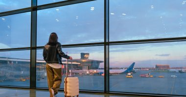 A young woman waits with her suitcase by an airport window as she prepares to board a flight. (Shutterstock Photo)
