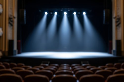 Empty theater stage with spotlights illuminating the center, rows of unoccupied seats visible in the foreground. (Shutterstock Photo)