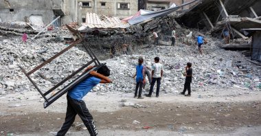 A boy walks carrying a table on his back past salvage workers scrapping the rubble of a building destroyed in Israel's war in Gaza, Palestine. (AFP Photo)