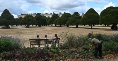 A gardener (R) works on the grounds as visitors (L) sit on a bench, looking out at the sun-scorched grass at Hampton Court, London, U.K., Aug. 20, 2026. (AFP Photo)