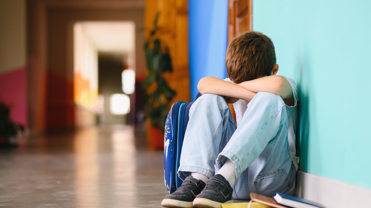 A boy is sitting in a school hallway with his hands on his knees and his head between his legs. (Shutterstock Photo)