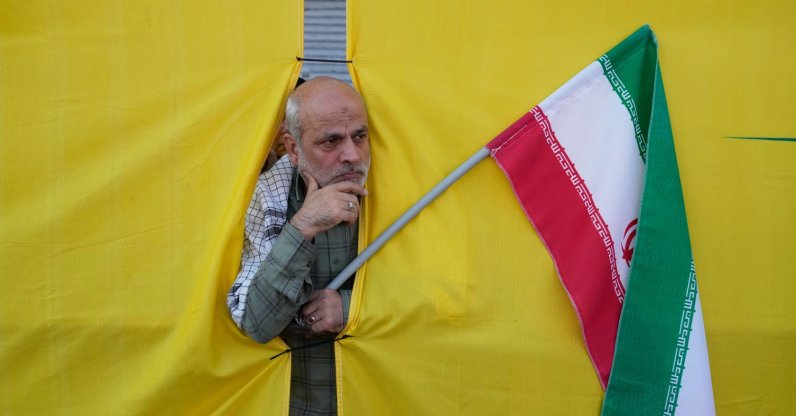 A man holding an Iranian flag peers through an opening in a curtain during the state-organized parade in Tehran, Iran, Sept. 18, 2026. (AP Photo)