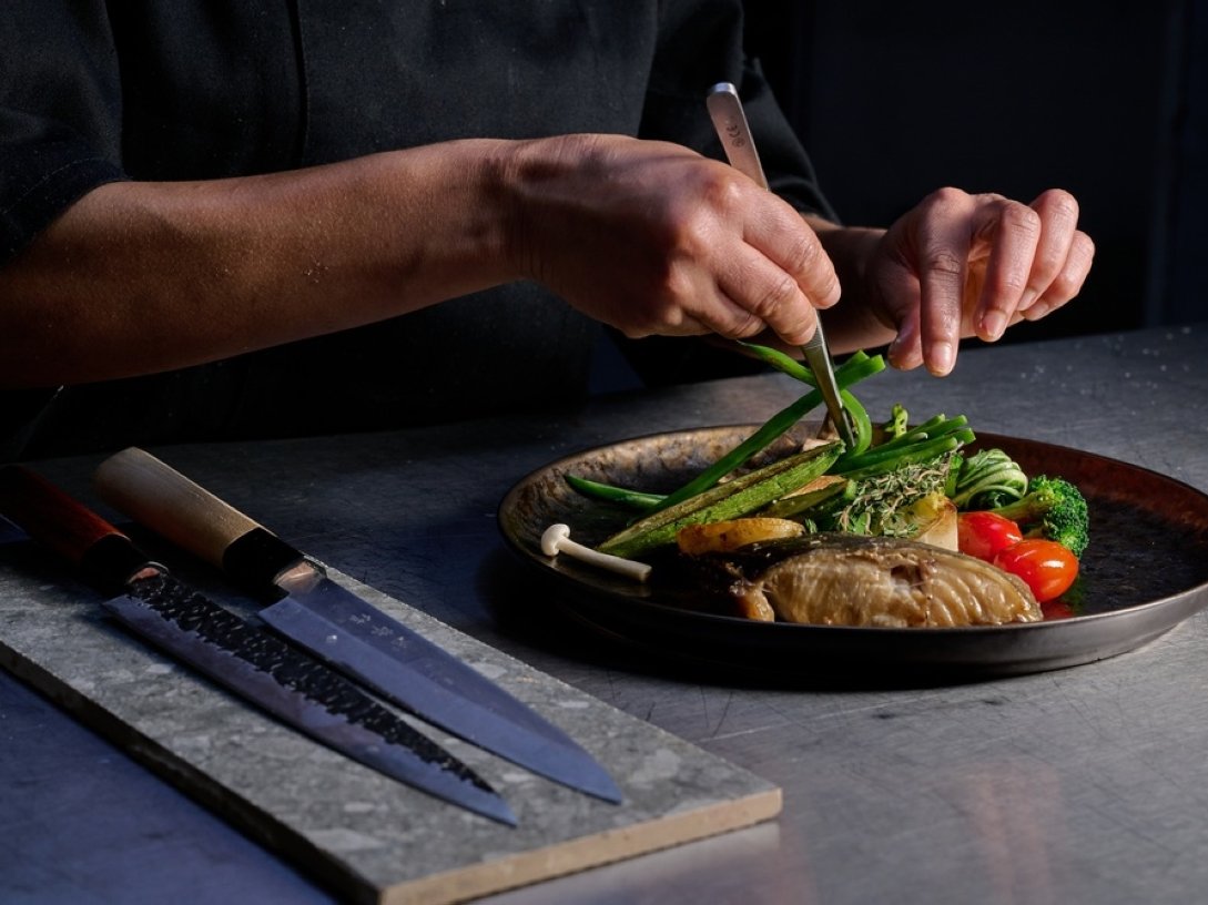 A professional chef meticulously plating a gourmet grilled fish dish with fresh seasonal vegetables using culinary tweezers. (Shutterstock Photo)