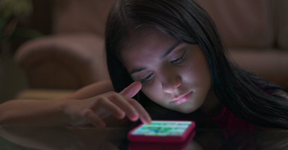 Teenage girl at a table engrossed in her phone, with glowing light illuminating her face, illustrating youth caught in social media addiction and tech disconnection. (Shutterstock Photo)