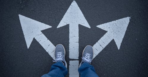 A person stands at a crossroads with multiple arrows pointing in different directions on the ground. (Shutterstock Photo)