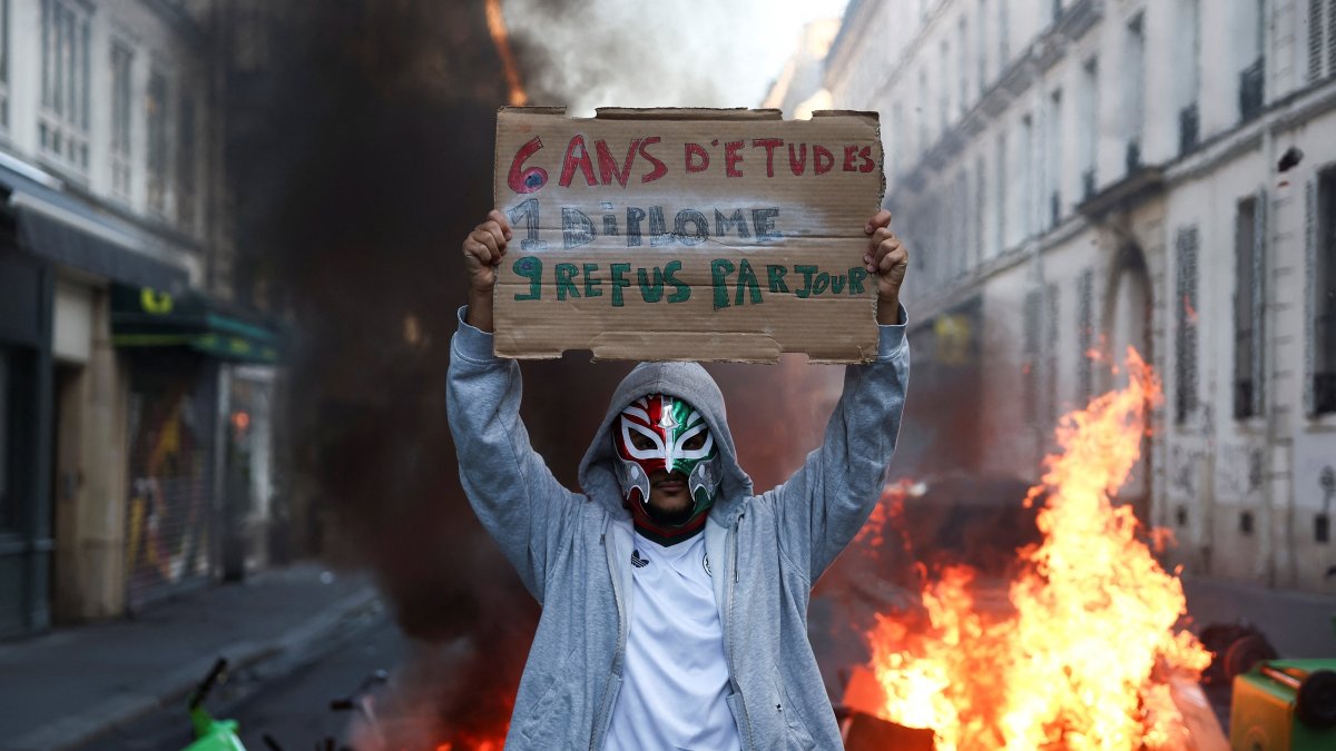 A person wearing a mask holds a placard reading "6 years of education, 1 diploma, 9 rejections a day" in front of burning flames, the day after a national day of demonstrations against the cost of living and rising fuel prices, Paris, France, Oct. 18, 2026. (Reuters Photo)