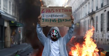 A person wearing a mask holds a placard reading "6 years of education, 1 diploma, 9 rejections a day" in front of burning flames, the day after a national day of demonstrations against the cost of living and rising fuel prices, Paris, France, Oct. 18, 2026. (Reuters Photo)