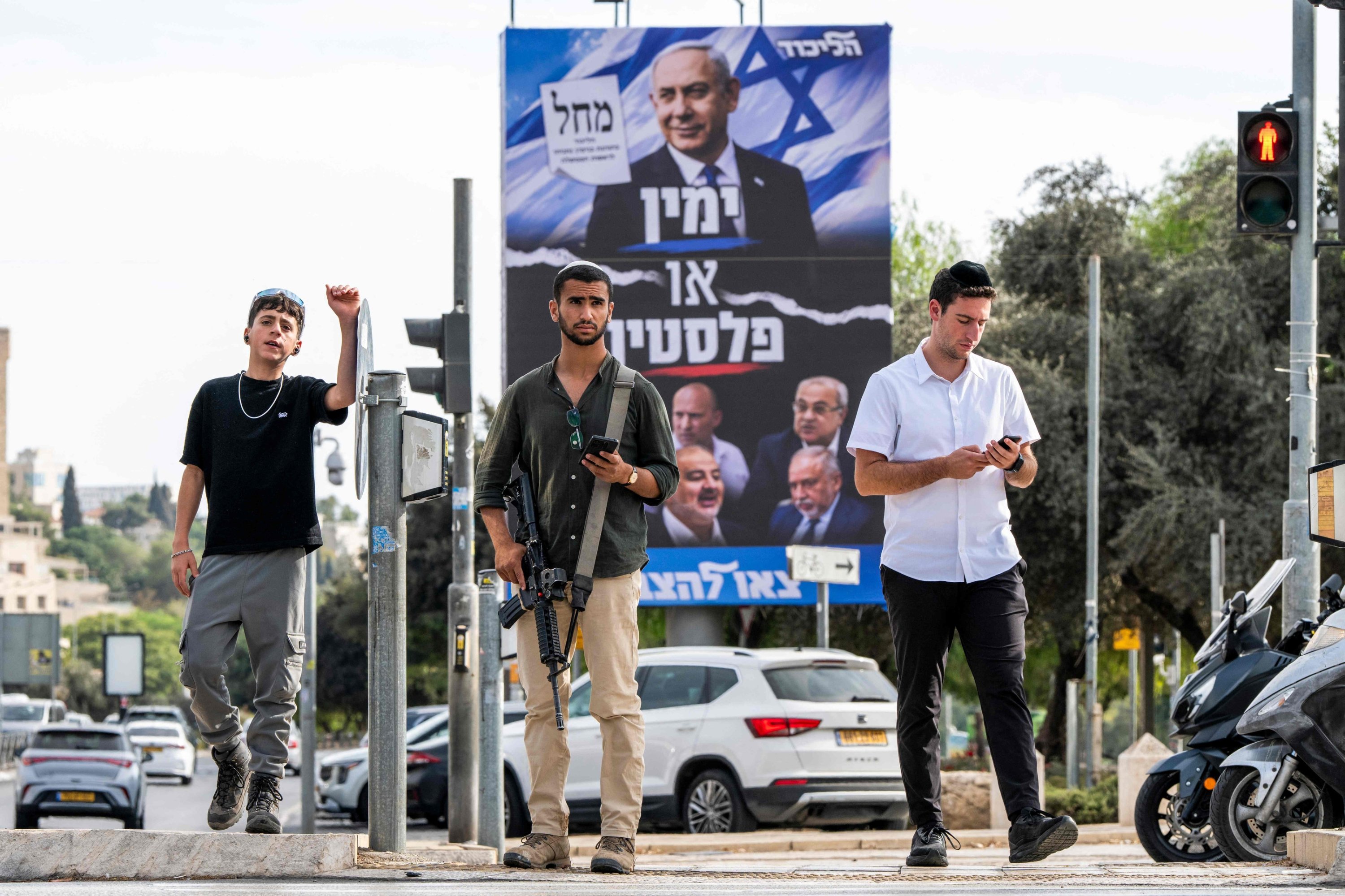 An armed Israeli man (C) waits with others to cross an intersection near an electoral campaign billboard for Israeli Prime Minister Benjamin Netanyahu's Likud party, west Jerusalem, Israel, Oct. 20, 2026. (AFP Photo)