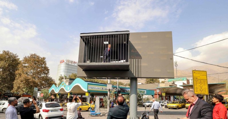 Pedestrians take pictures of an Iranian actor wearing a mask depicting U.S. President Donald Trump as he poses inside a mock prison cell installed onto a street billboard at Tajrish Square in Tehran, Oct. 21, 2026. (AFP Photo)